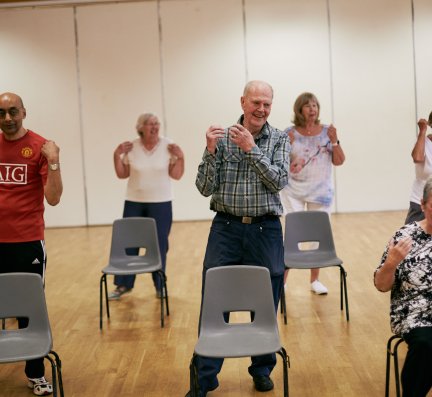 a small group of older people in a community hall wearing casual clothes. They are smiling and sitting or standing to do gentle exercises and movements.