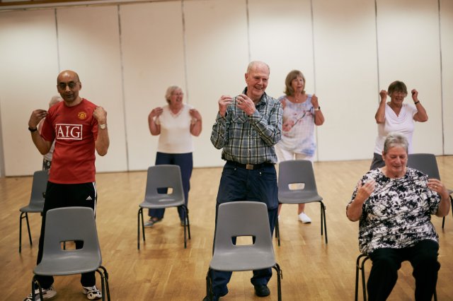 a small group of older people in a community hall wearing casual clothes. They are smiling and sitting or standing to do gentle exercises and movements.