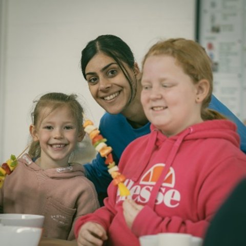 an image that shows a smiling teacher with two school age girls, sat at activity tables and making fruit kebabs