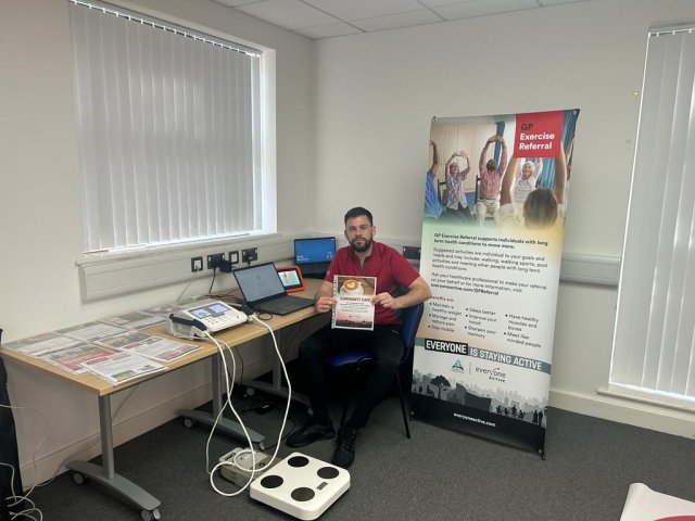 an image of a casually dressed male healthcare professional sat in a consulting room next to a large, colourful banner that promotes physical activity for improved patient health